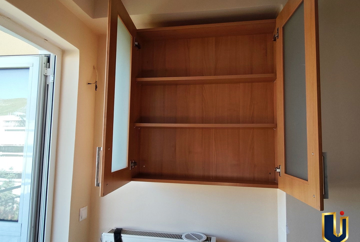 Open wooden kitchen cupboard with shelves beside a balcony door in a Paiania apartment