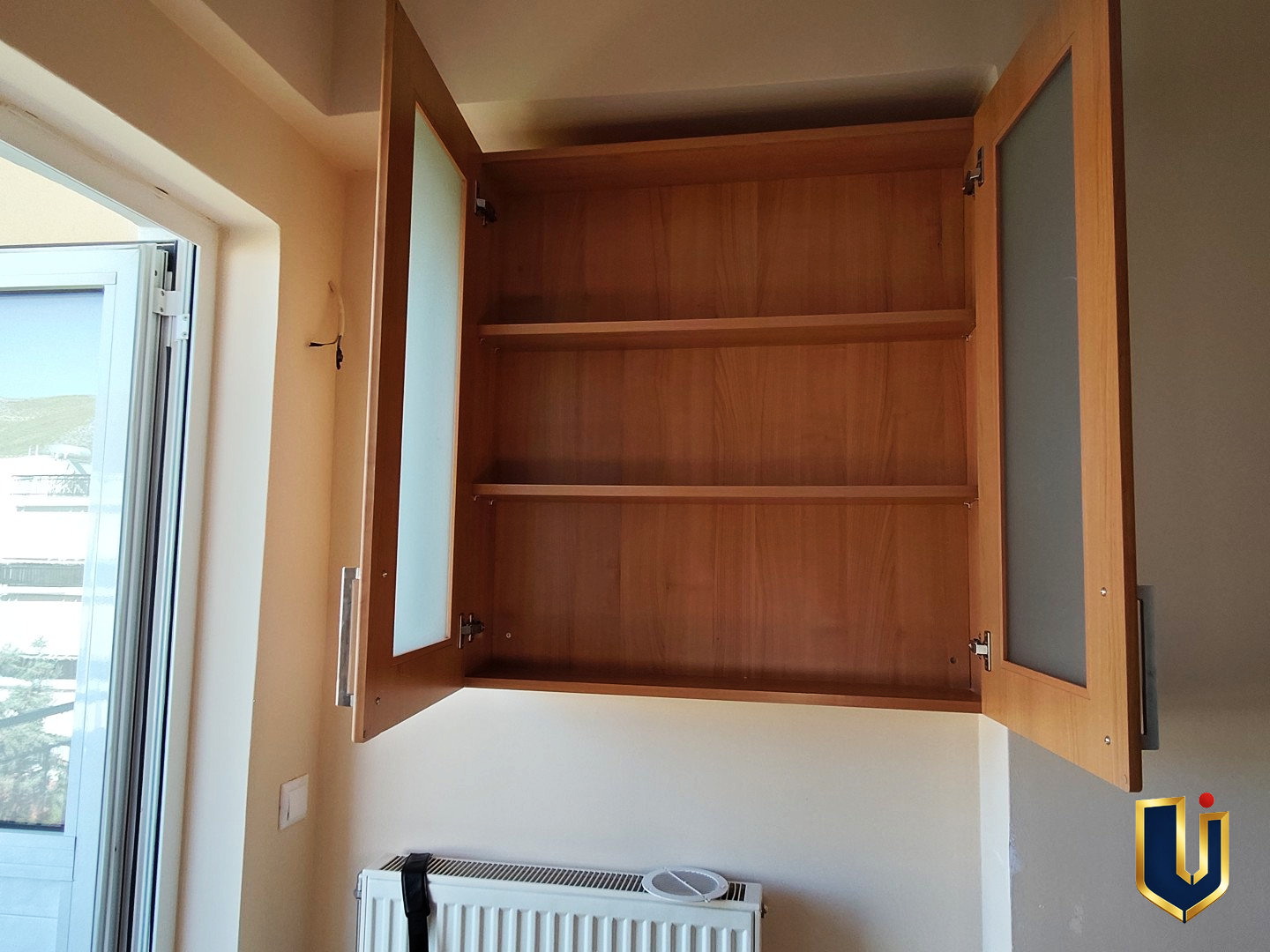 Open wooden kitchen cupboard with shelves beside a balcony door in a Paiania apartment