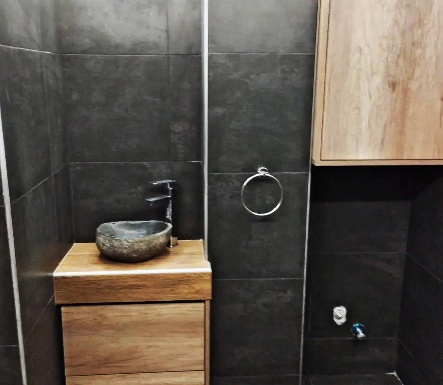 Bathroom with a countertop basin, wood-finish vanity and dark tiled walls in Ano Patisia, Athens