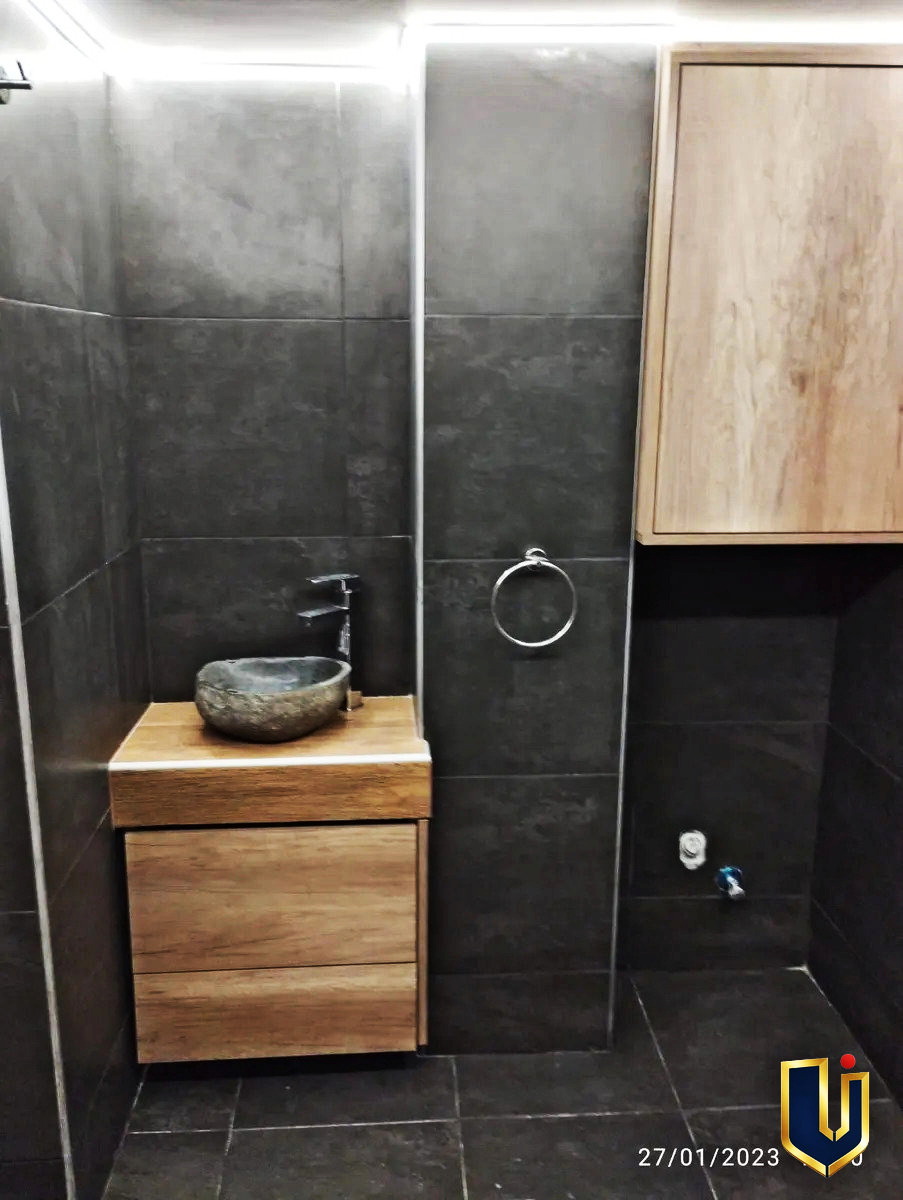 Bathroom with a countertop basin, wood-finish vanity and dark tiled walls in Ano Patisia, Athens