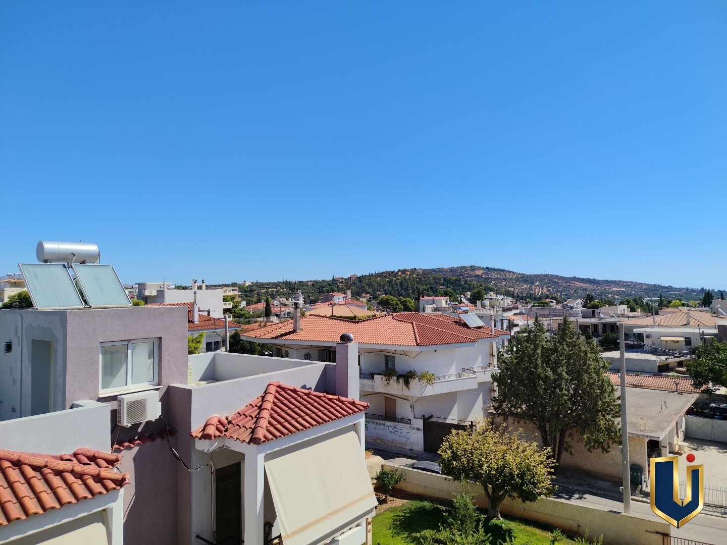View from a Paiania apartment bedroom over neighbouring homes, tiled rooftops and trees towards a distant hill